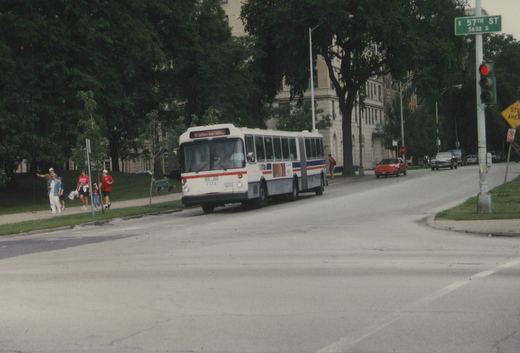 A white CTA articulated bus with the number 7174 is turning a corner on a tree-lined city street, with pedestrians on the sidewalk and cars in the background. A street sign reads 'E 57TH ST'.