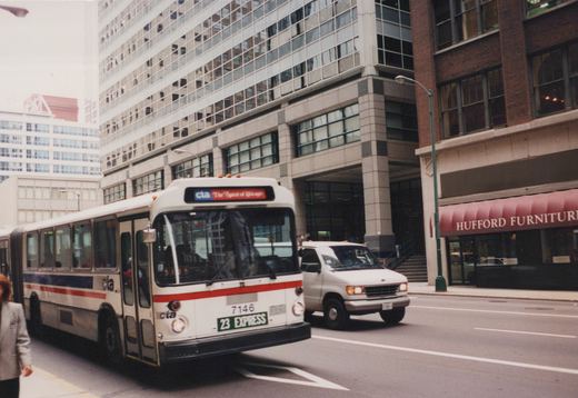 A white CTA bus with red and blue stripes, numbered 7146, is driving down a city street past a building with a Hufford Furniture awning.