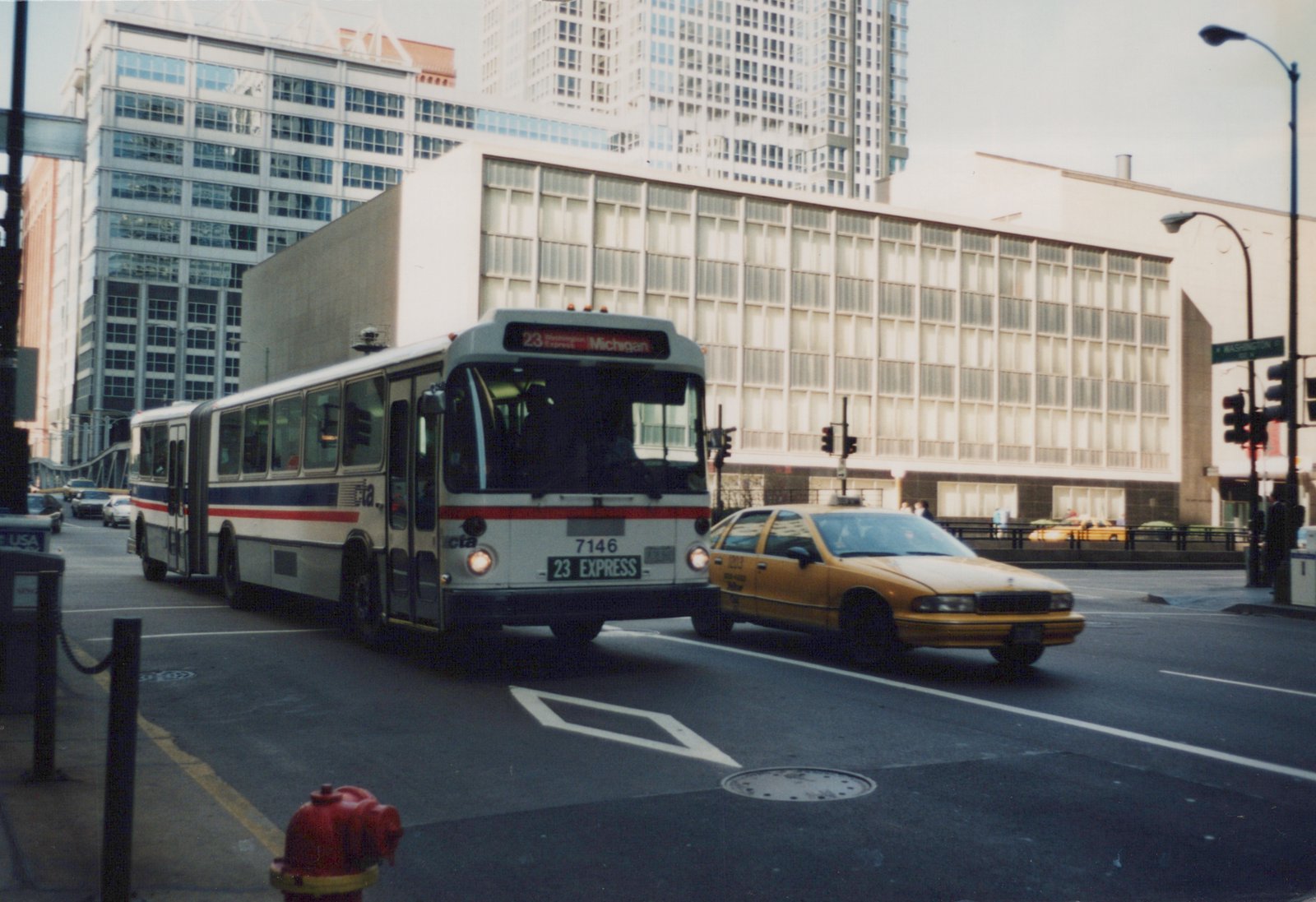 A white articulated bus with red and blue stripes, marked CTA 7146 and 23 EXPRESS, turns at an intersection with a yellow taxi cab nearby, surrounded by tall buildings on a cloudy day.