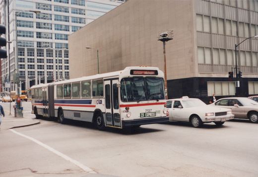 A white and blue articulated bus with the number 7137 and route 23 Express is turning on a city street, surrounded by cars and tall buildings.
