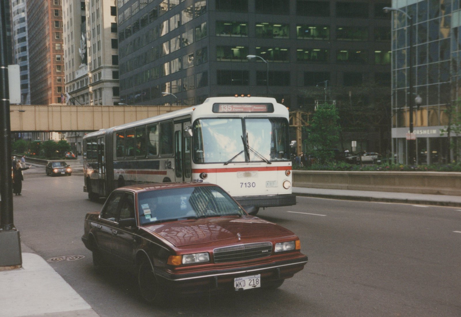 A white and red Chicago Transit Authority bus with the number 7130 and route 135 on its sign is driving on a city street, with a red car in the foreground and tall buildings in the background.