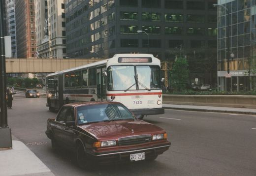 A white and red Chicago Transit Authority bus with the number 7130 and route 135 on its sign is driving on a city street, with a red car in the foreground and tall buildings in the background.