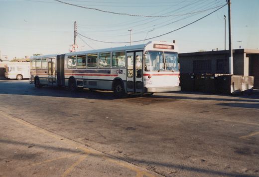 A white, red, and blue articulated bus with the number 7130 on its front, parked in a paved lot with a building and dumpsters in the background.