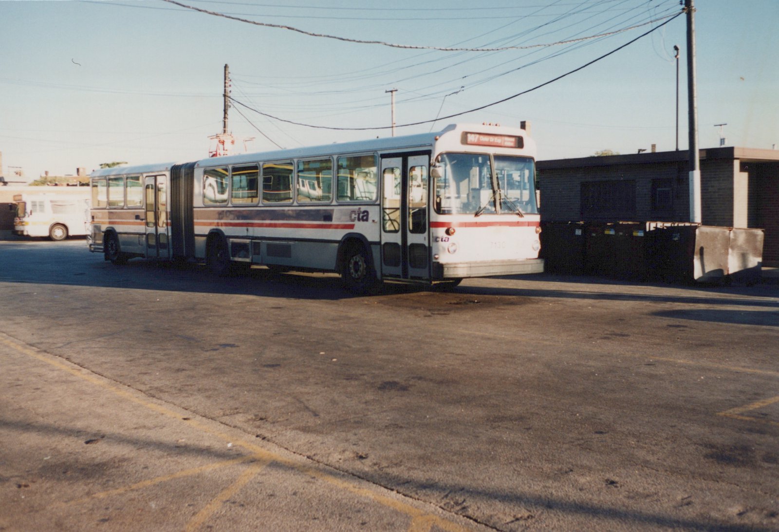 A white, red, and blue articulated bus with the number 7130 on its front, parked in a paved lot with a building and dumpsters in the background.