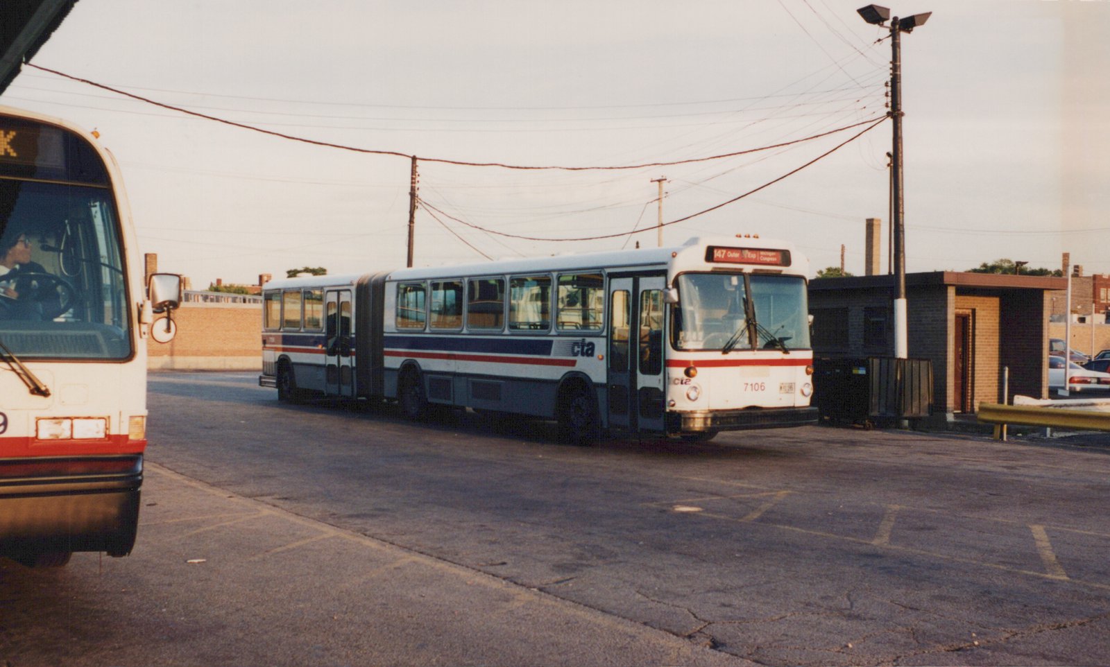 A white, red and blue articulated CTA bus, number 7106, is parked on an asphalt lot with another bus partially visible to the left and a small building in the background.