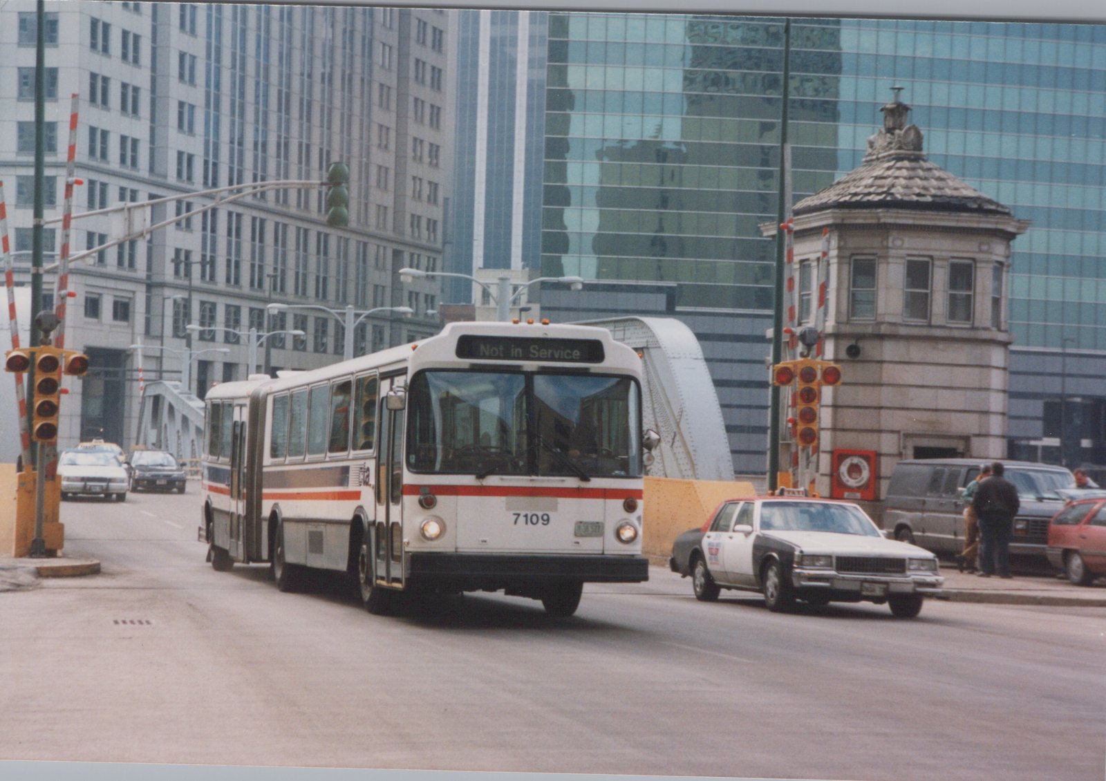 An articulated Chicago Transit Authority bus numbered 7109, displaying 'Not in Service' on its front sign, drives across a street bridge in a city with modern buildings, alongside a taxi and other cars.