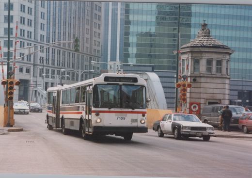 An articulated Chicago Transit Authority bus numbered 7109, displaying 'Not in Service' on its front sign, drives across a street bridge in a city with modern buildings, alongside a taxi and other cars.