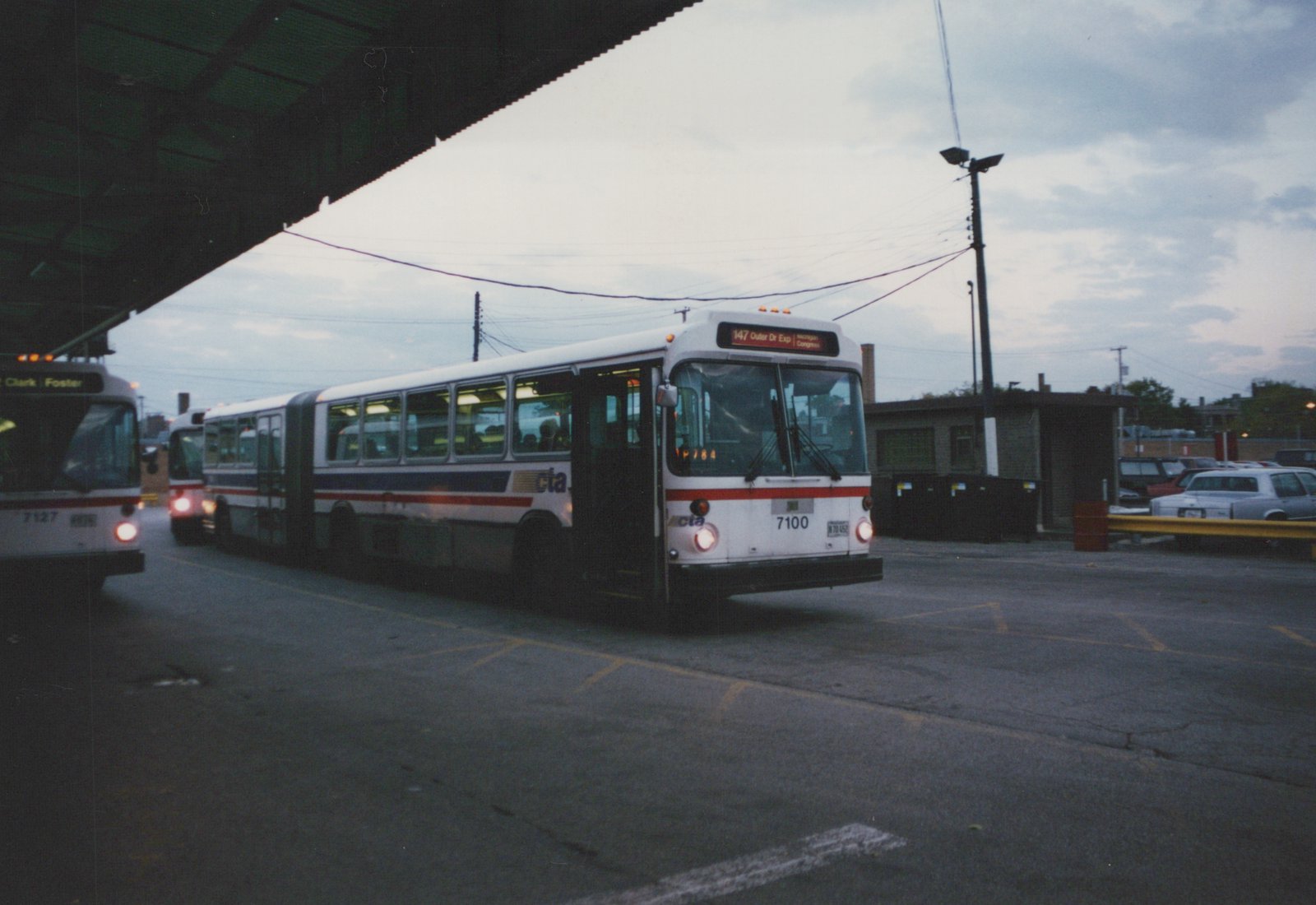 A white CTA articulated bus with the number 7100 is parked at a terminal, with other buses and cars in the background under an overcast sky.