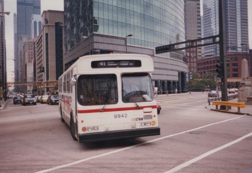 A white CTA bus with a red stripe, numbered 9942, is driving on a city street with tall buildings and other traffic around it.