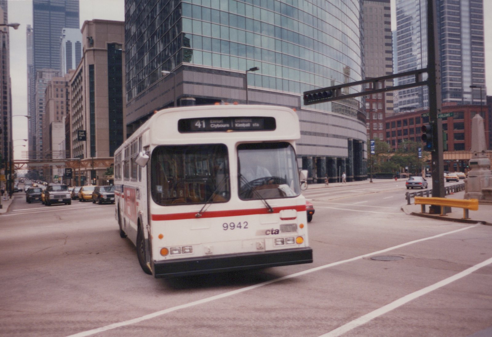 A white CTA bus with a red stripe, numbered 9942, is driving on a city street with tall buildings and other traffic around it.