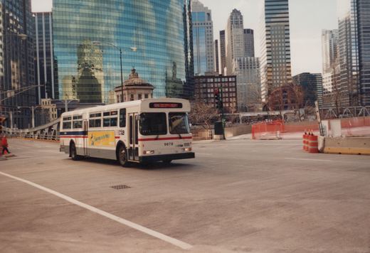 A white CTA bus numbered 9878, with a blue and red stripe, is turning on a city street with tall glass buildings behind it on an overcast winter day.