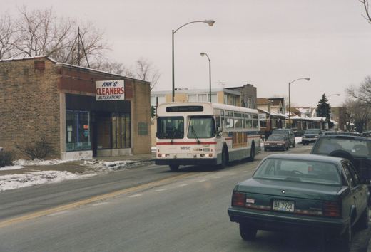 A white and red CTA bus with the number 9850 on its front, driving down a street lined with snow and parked cars. A green car is in the foreground with a visible license plate, and a brick building with a 'Ann's Cleaners' sign is on the left.