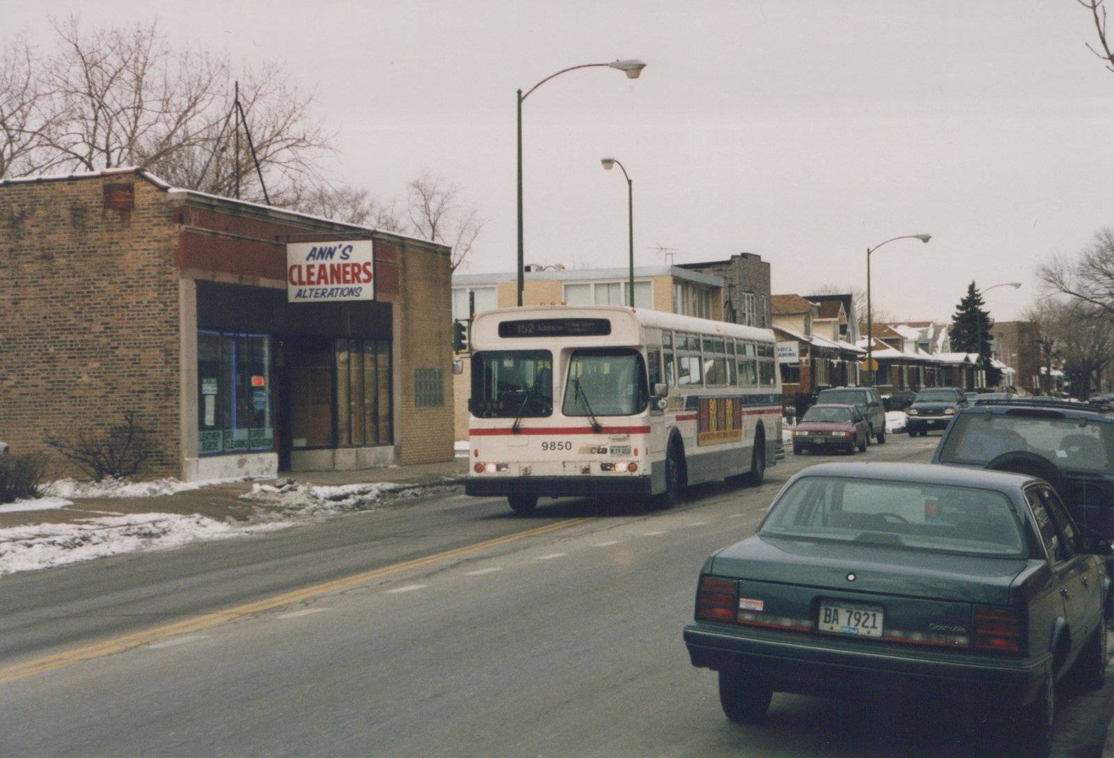 A white and red CTA bus with the number 9850 on its front, driving down a street lined with snow and parked cars. A green car is in the foreground with a visible license plate, and a brick building with a 'Ann's Cleaners' sign is on the left.