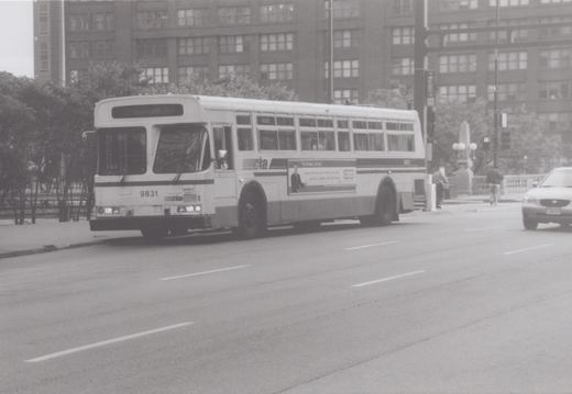 A black and white photo shows a CTA bus, number 9831, at an intersection with a late 1990s sedan and two pedestrians nearby.