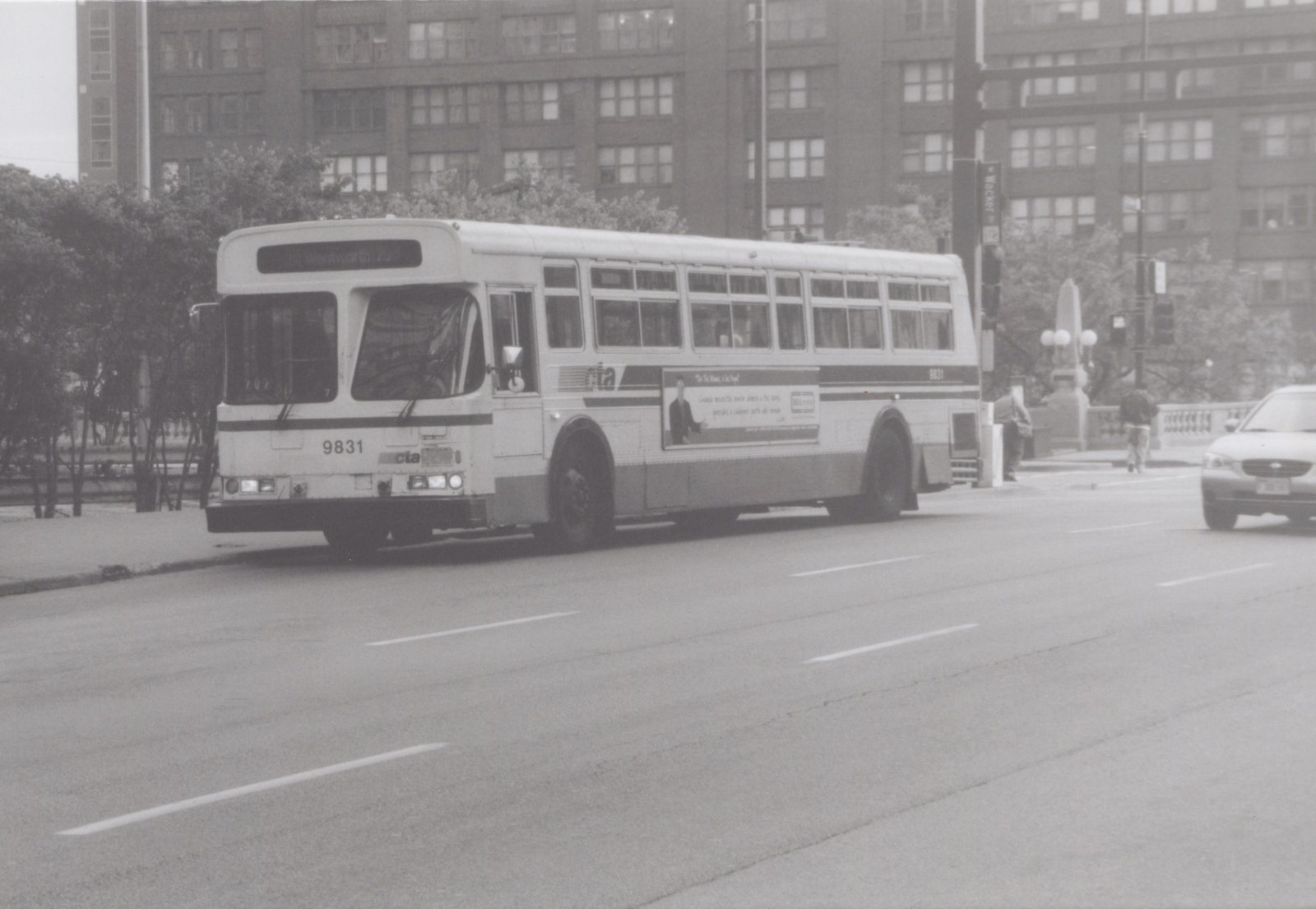 A black and white photo shows a CTA bus, number 9831, at an intersection with a late 1990s sedan and two pedestrians nearby.