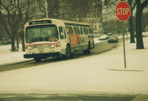 A white Chicago Transit Authority bus with a red stripe, number 9293, drives on a snowy street past a red stop sign, with bare trees and a brick building in the background.