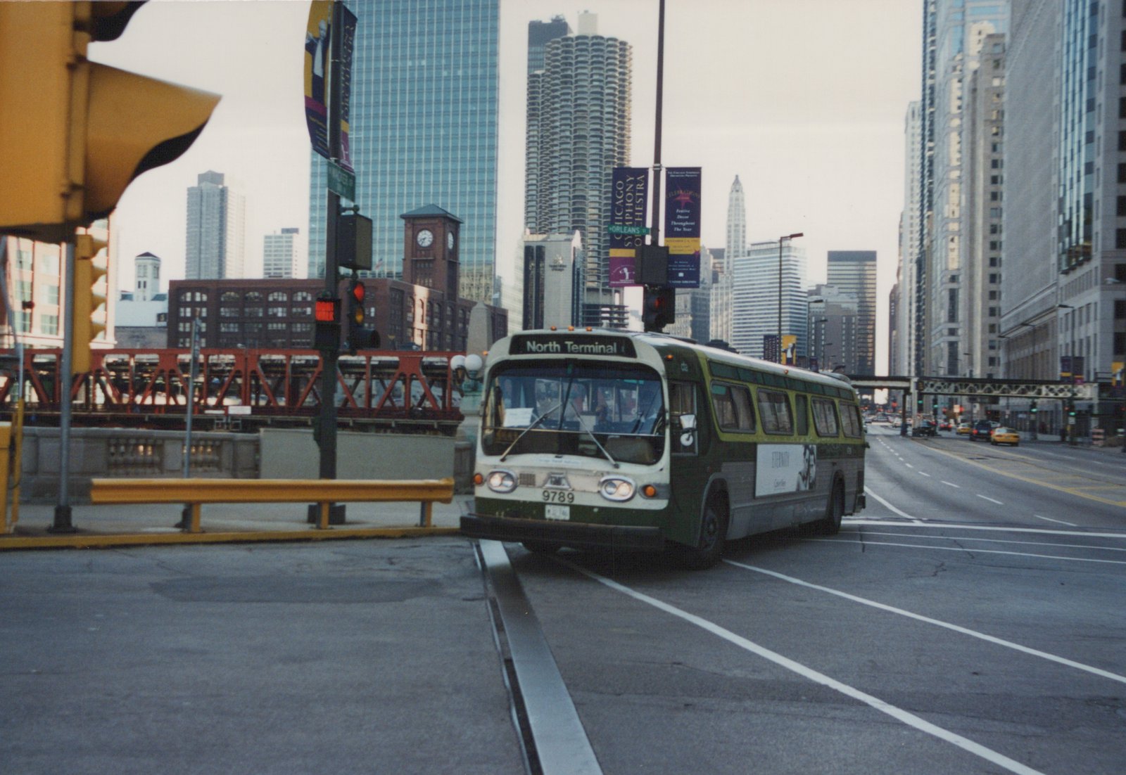 A green and white CTA bus with the number 9789 and destination sign reading 'North Terminal' is stopped at an intersection in a city with tall buildings and elevated train tracks in the background.