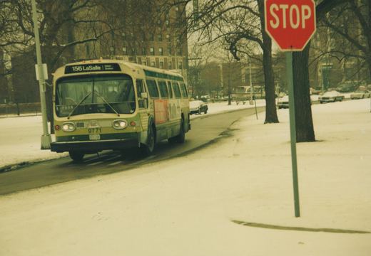A vintage CTA bus, number 9771, drives down a snow-covered street in winter with a stop sign visible on the right and bare trees lining the road.