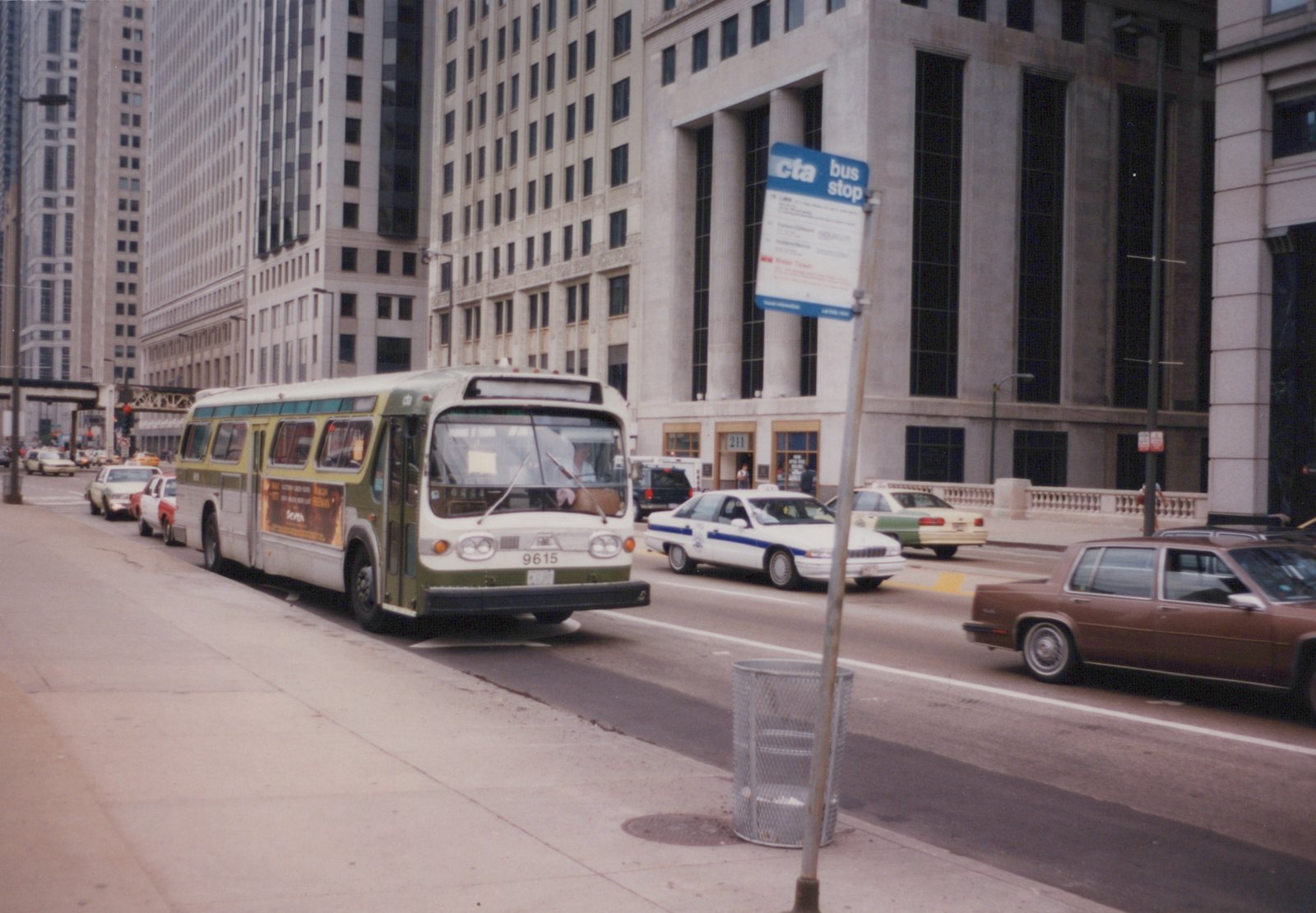 A green and white Chicago Transit Authority bus numbered 9615 is driving on a city street with tall buildings, a police car, and other vehicles nearby.