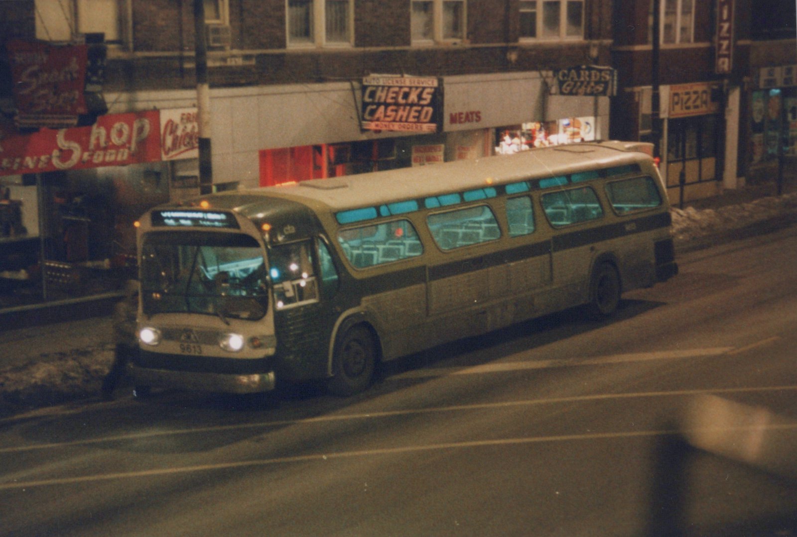 A white and dark green CTA bus, number 9613, is parked on a snowy city street at night, with signs for a check cashing service and a pizza place visible behind it.