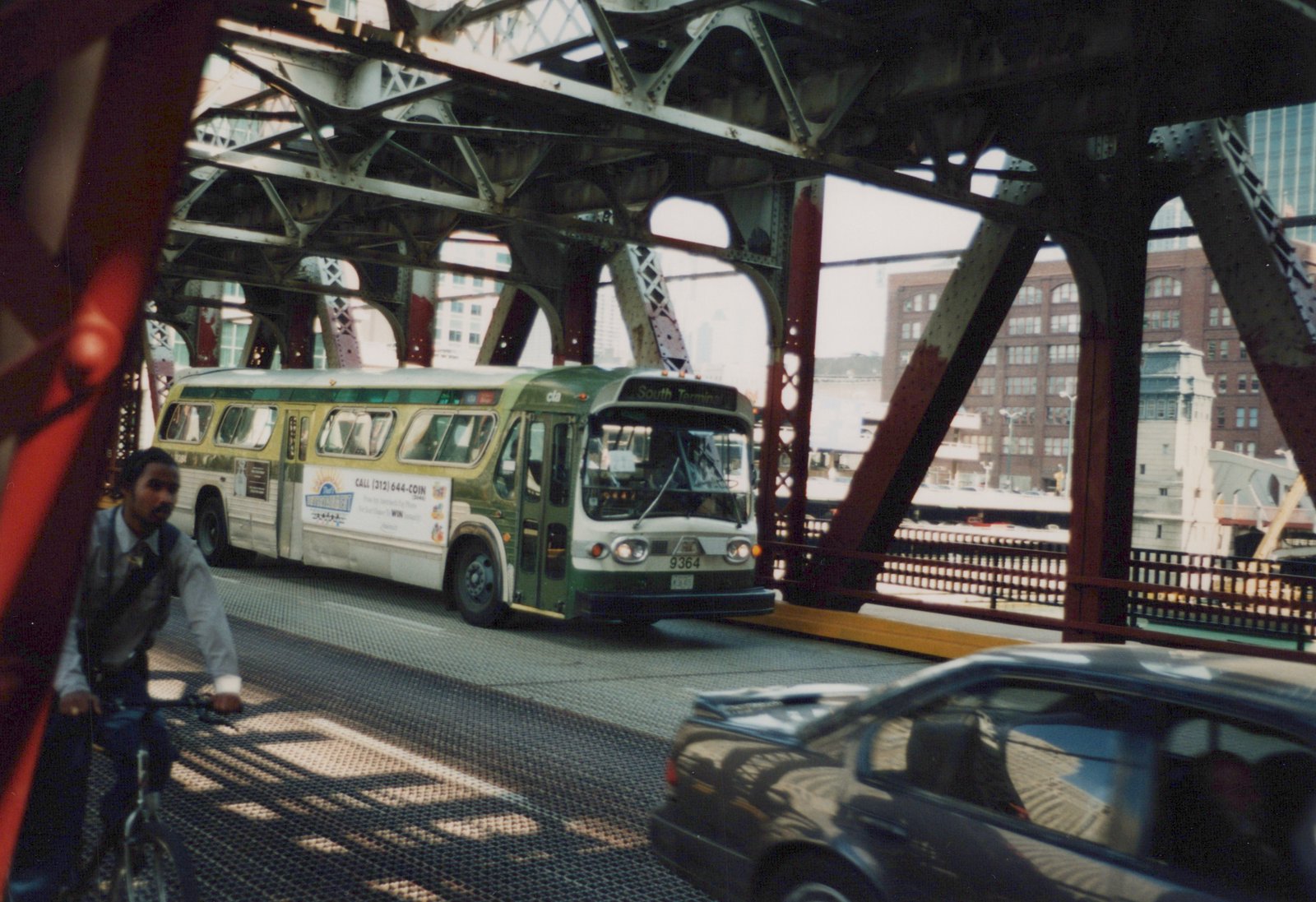 A green and white CTA bus, number 9364, is on a metal truss bridge with a 'South Terminal' destination sign, accompanied by a cyclist and a dark car.