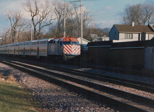 A red, white, and blue Metra locomotive numbered 154 pulls a silver train of gallery cars along tracks beside a grassy embankment and houses, with bare trees in the background under a clear sky.