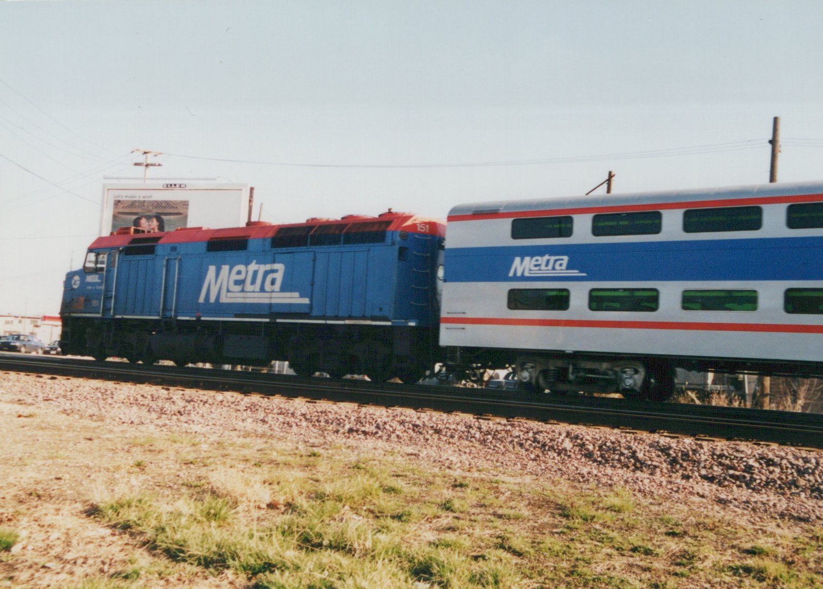 A blue and red Metra locomotive numbered 151 pulls a white and blue double-decker commuter car on a railroad track with a grassy foreground and a billboard in the background.