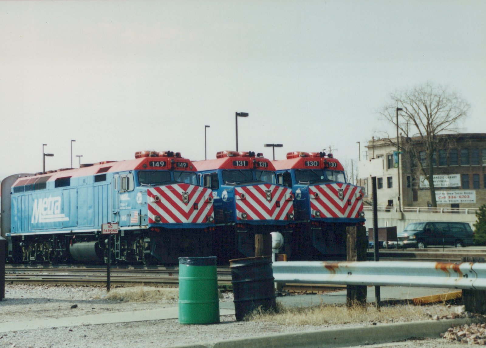 Three blue and red Metra locomotives with the numbers 149, 131, and 130 are parked side by side in a gravel yard, with a building and a van visible in the background.