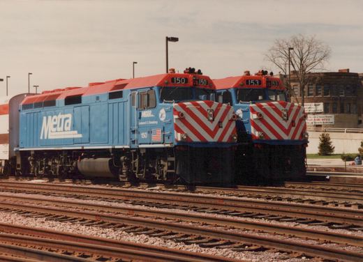 A roster shot of two blue and red Metra locomotives, numbers 150 and 153, parked on parallel tracks in a yard with bare trees in the background.