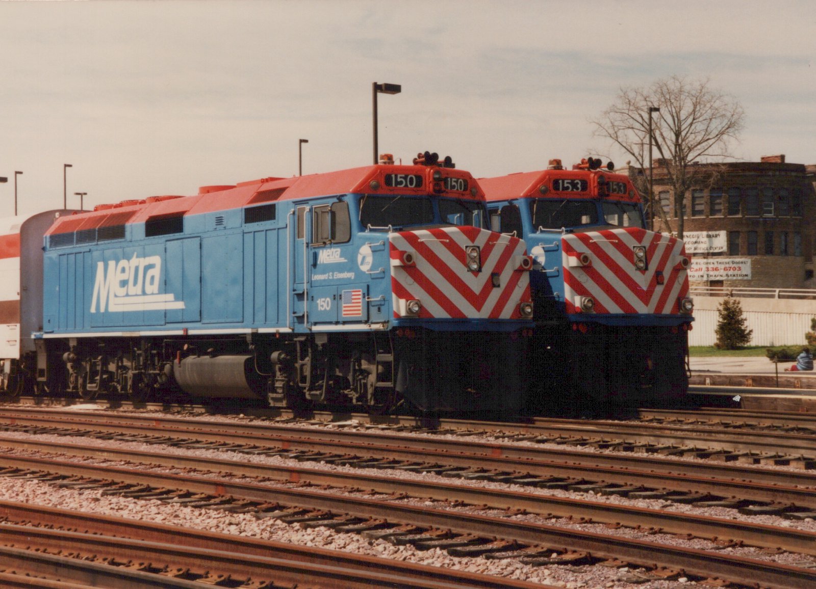 A roster shot of two blue and red Metra locomotives, numbers 150 and 153, parked on parallel tracks in a yard with bare trees in the background.