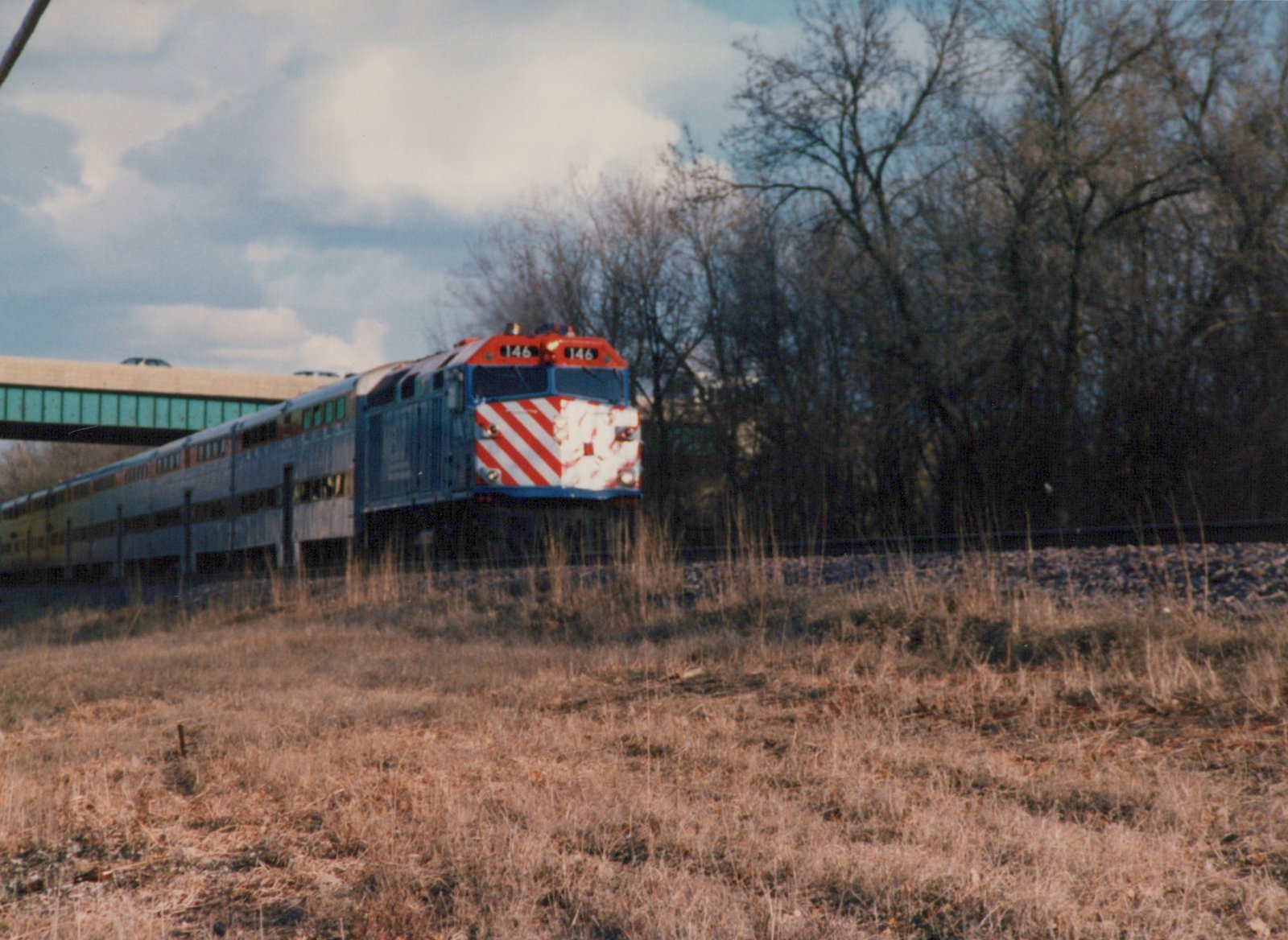 A blue, red, and white Metra train with locomotive 146 leads a line of double-decker commuter cars along a track near a bridge, with bare trees and dry grass under an overcast sky.