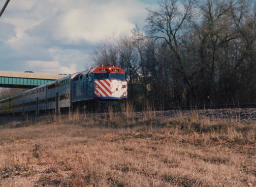 A blue, red, and white Metra train with locomotive 146 leads a line of double-decker commuter cars along a track near a bridge, with bare trees and dry grass under an overcast sky.