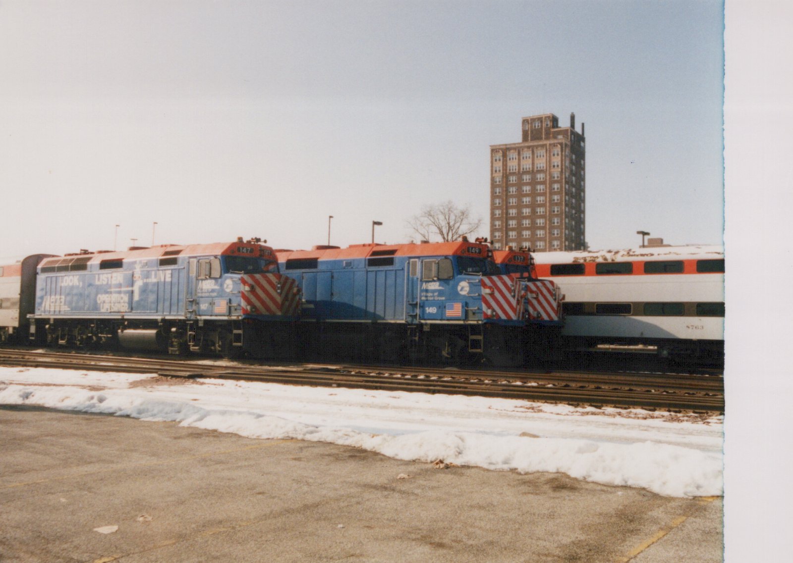 Two blue and orange Metra locomotives, numbers 147 and 149, coupled to a white and orange passenger car on a snowy railroad yard with a tall building in the background.