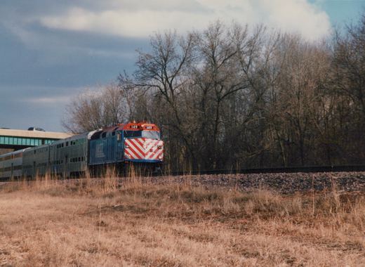 A blue Metra locomotive with red and white chevron stripes, number 149, pulls a line of gray double-decker passenger cars on a track beside a grassy embankment with bare trees in the background under an overcast sky.