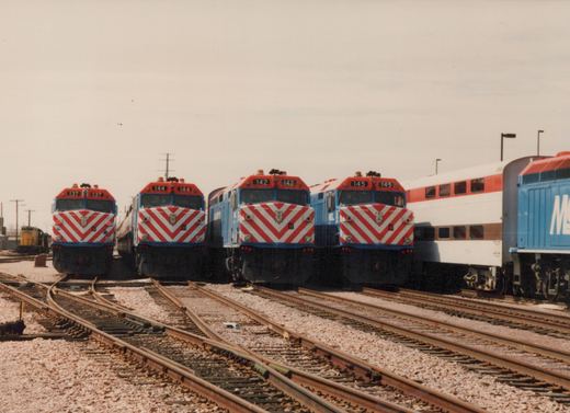 A row of four blue, white, and red Metra diesel locomotives with numbers 137, 144, 142, and 145, parked on multiple tracks in a gravel yard with a double-decker passenger car behind them.