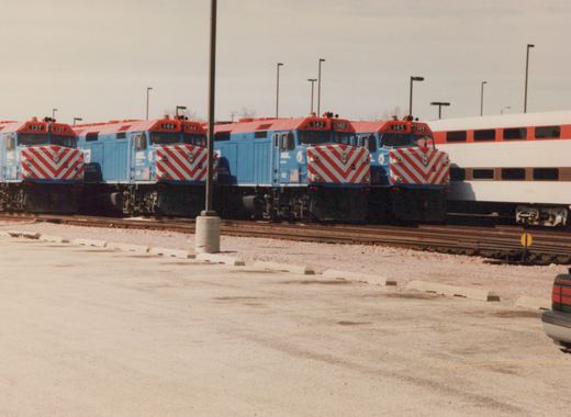 A row of four blue and red Metra locomotives, numbers 137, 144, 142, and 145, are parked on tracks in a rail yard under an overcast sky, with a white Amtrak passenger car partially visible behind them.