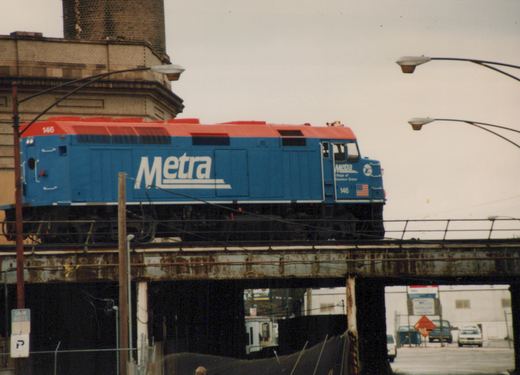 A blue and red Metra diesel locomotive numbered 146 travels on an elevated steel bridge over a street in a winter scene.