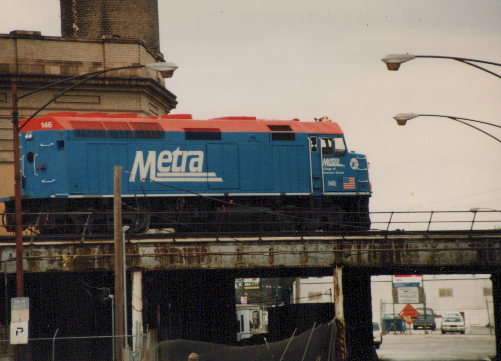 A blue and red Metra diesel locomotive numbered 146 travels on an elevated steel bridge over a street in a winter scene.