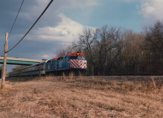 A red, white and blue Metra train with locomotive number 145 travels on a track beside a dry, grassy field with bare trees in the background under a cloudy sky.
