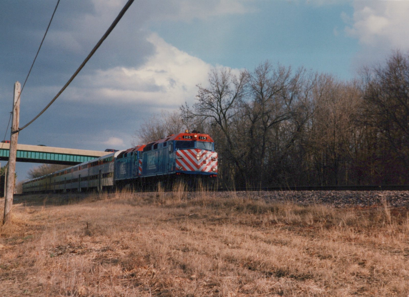 A red, white and blue Metra train with locomotive number 145 travels on a track beside a dry, grassy field with bare trees in the background under a cloudy sky.