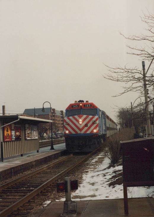 A red, white, and blue Metra locomotive numbered 145 pulls a train of passenger cars through a station with snow on the ground and bare trees in the background.