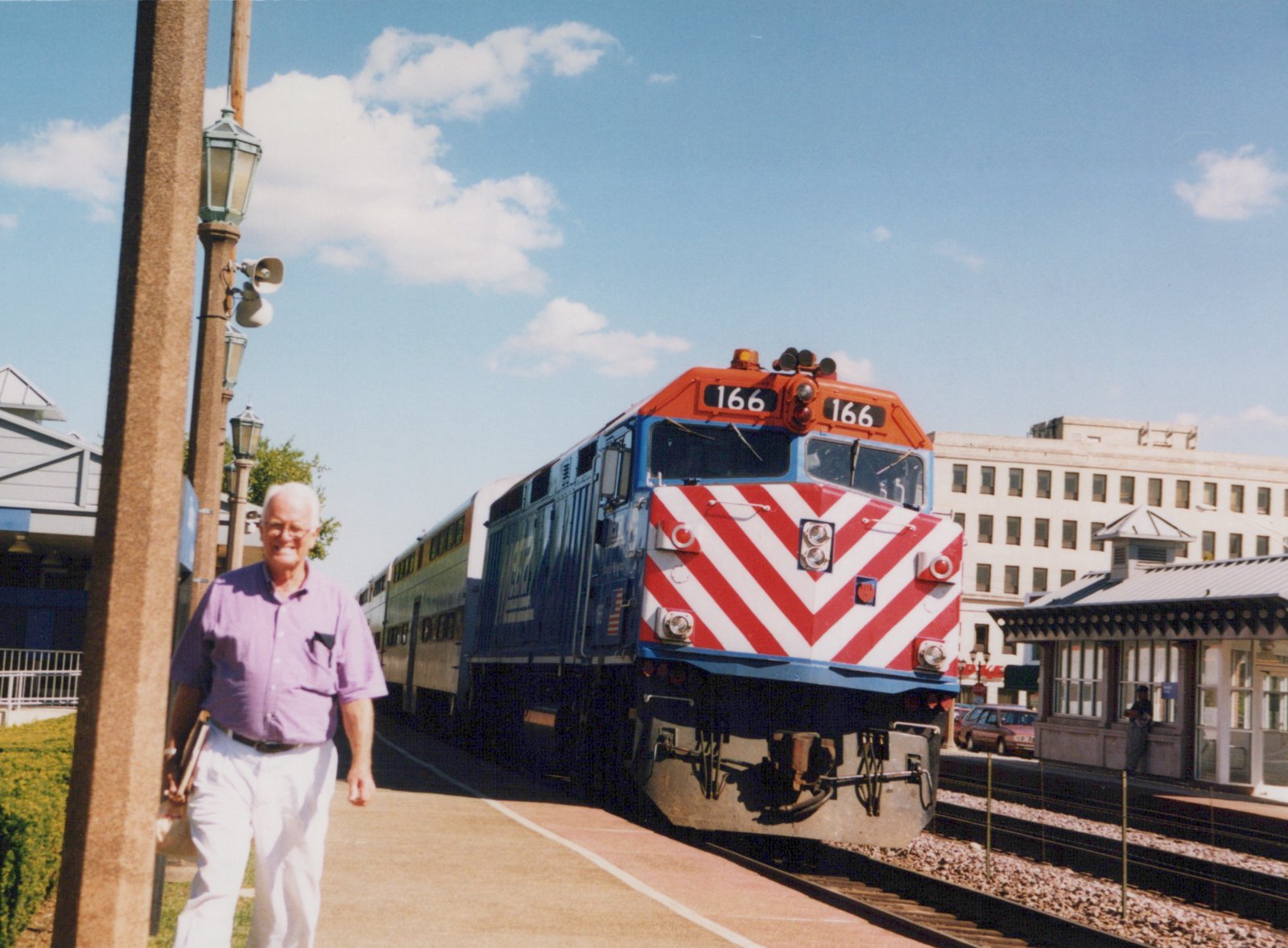 A blue and red Metra locomotive, number 166, sits on the tracks next to a station platform under a clear blue sky; a man in a purple shirt walks on the platform in the foreground.