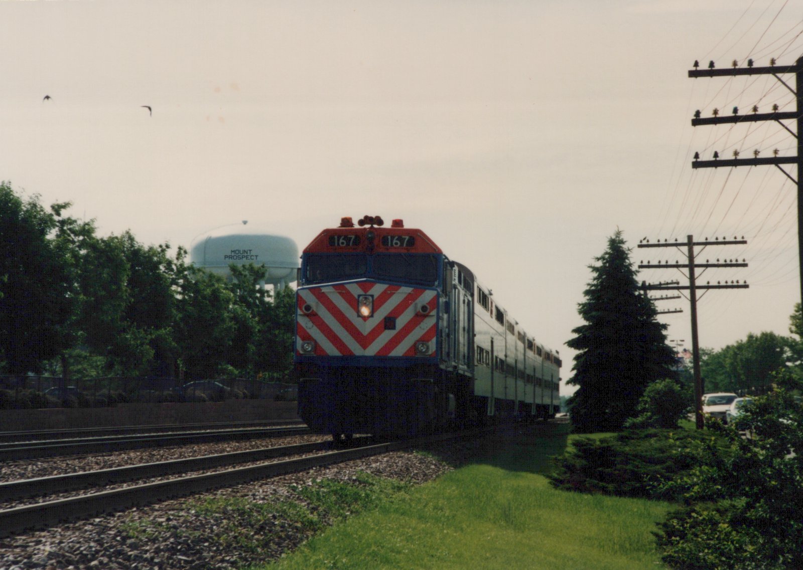 A Metra train with a red, white and blue locomotive numbered 167 is traveling on a set of tracks with a water tower in the background that says 'MOUNT PROSPECT'.