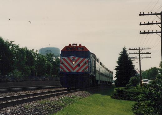 A Metra train with a red, white and blue locomotive numbered 167 is traveling on a set of tracks with a water tower in the background that says 'MOUNT PROSPECT'.