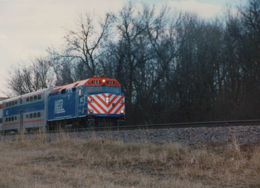 A blue Metra locomotive with red and white chevron stripes, numbered 168, pulls double-decker passenger cars along tracks bordered by dry grass and bare trees under an overcast sky.