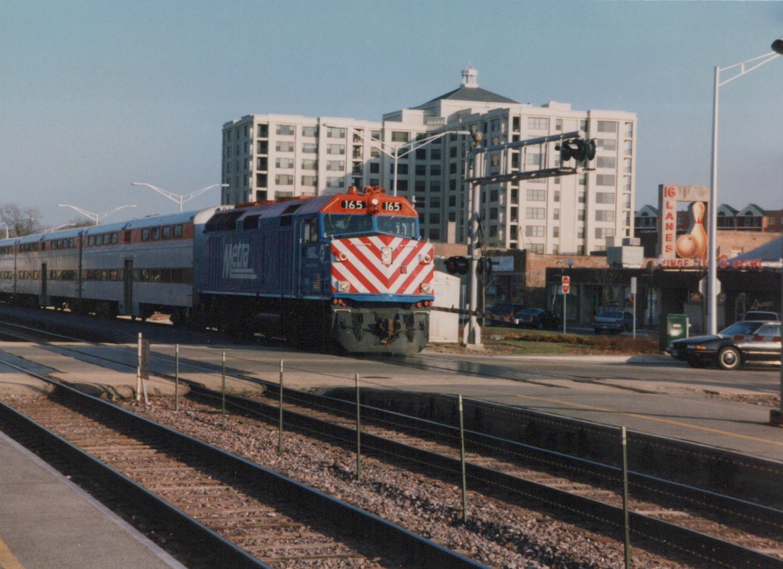 A blue and red Metra locomotive numbered 165 is pulling silver gallery cars across a street at a grade crossing, with a multi-story building and a bowling alley sign in the background.