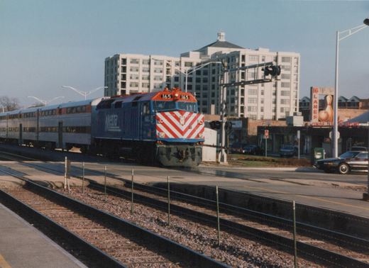 A blue and red Metra locomotive numbered 165 is pulling silver gallery cars across a street at a grade crossing, with a multi-story building and a bowling alley sign in the background.