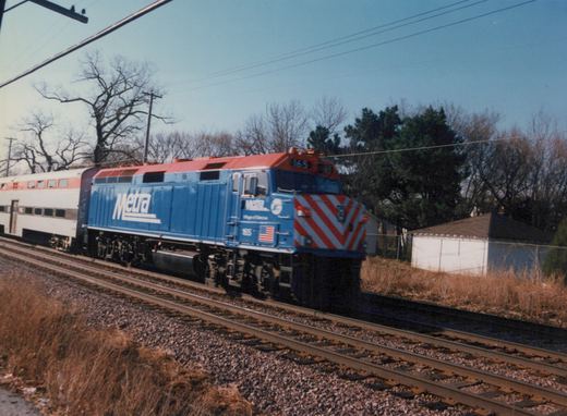 A blue and red Metra locomotive with the number 165 travels on tracks beside dry grass and bare trees under a clear blue sky.