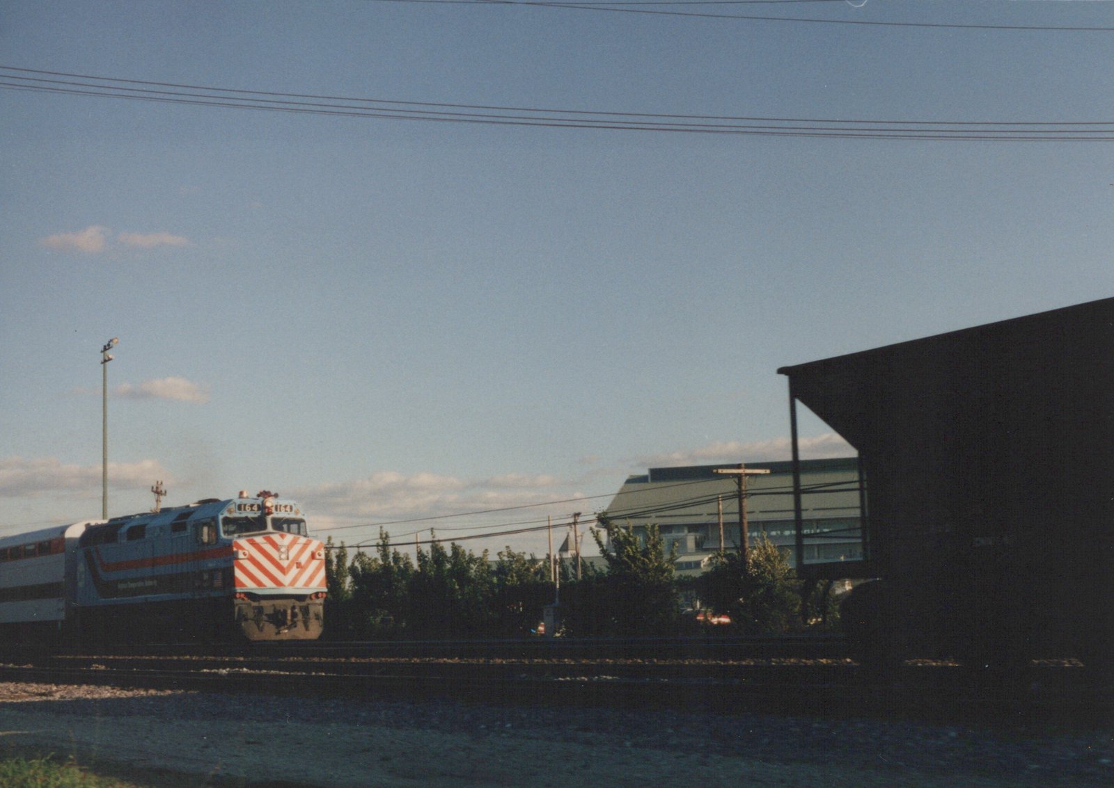 A Metra diesel locomotive numbered 164, painted in blue with red and white stripes, is shown pulling a double-decker passenger car on a sunny day alongside a building and trees.
