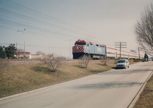 A Metra train with a blue locomotive and white passenger cars travels on railroad tracks beside a road with a white minivan, set against a backdrop of bare trees and a clear sky.